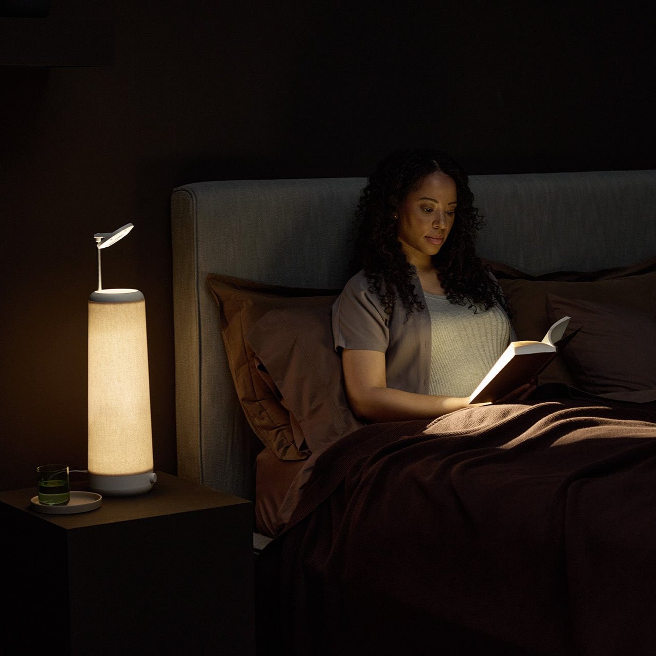 A woman lounges in bed, absorbed in her book under the soothing warm-white LED light of the NS01 Personal Task Lamp by Nightside. The room exudes calmness, dimly lit with brown sheets and a gray headboard for a cozy ambiance.