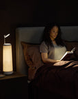 A woman lounges in bed, absorbed in her book under the soothing warm-white LED light of the NS01 Personal Task Lamp by Nightside. The room exudes calmness, dimly lit with brown sheets and a gray headboard for a cozy ambiance.