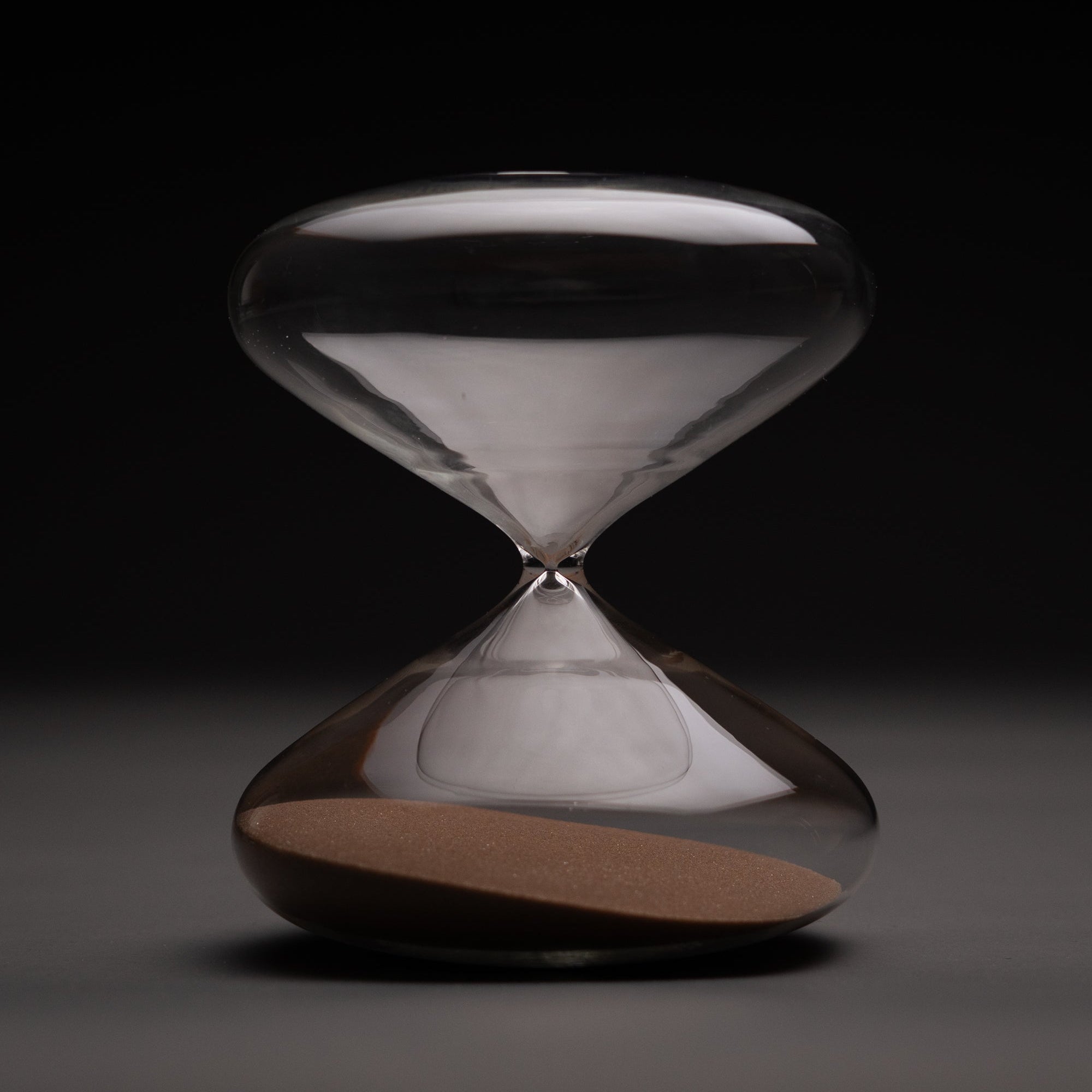The Intelligent Change Mindful Focus 30 Minute Hourglass, featuring clear glass and brown sand mostly in the bottom half, stands centered and well-lit against a dark background—ideal for illustrating time management.