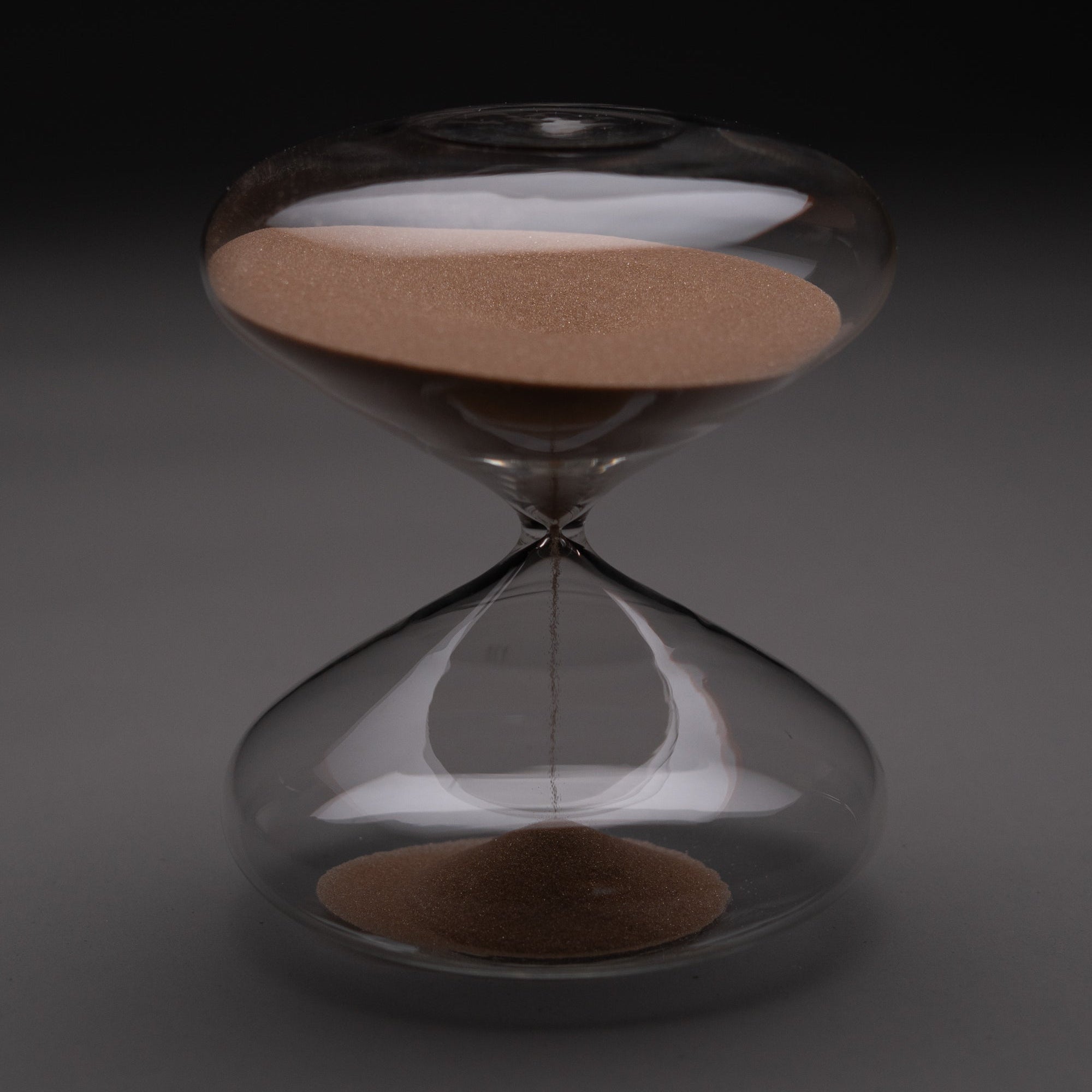 The Intelligent Change Mindful Focus 30 Minute Hourglass, featuring brown sand mostly in the upper half, stands upright on a gray surface with a dark background, representing mindfulness and effective time management.