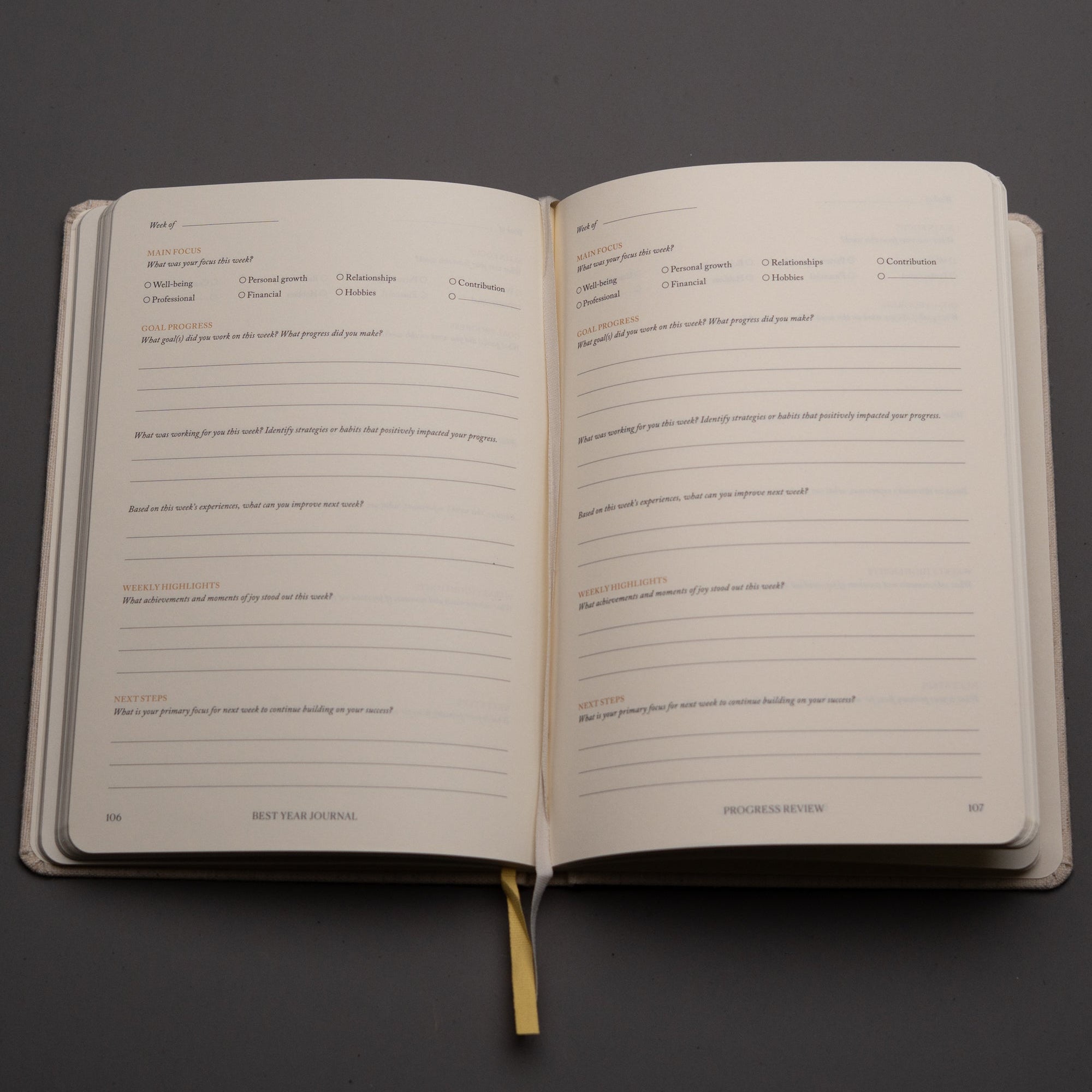 The Best Year Journal by Intelligent Change is open to cream-colored pages with self-reflection and goal review prompts. A yellow ribbon bookmark accents the inspiring planner, which rests on a gray surface for mindful, intentional living.