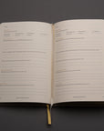 The Best Year Journal by Intelligent Change is open to cream-colored pages with self-reflection and goal review prompts. A yellow ribbon bookmark accents the inspiring planner, which rests on a gray surface for mindful, intentional living.