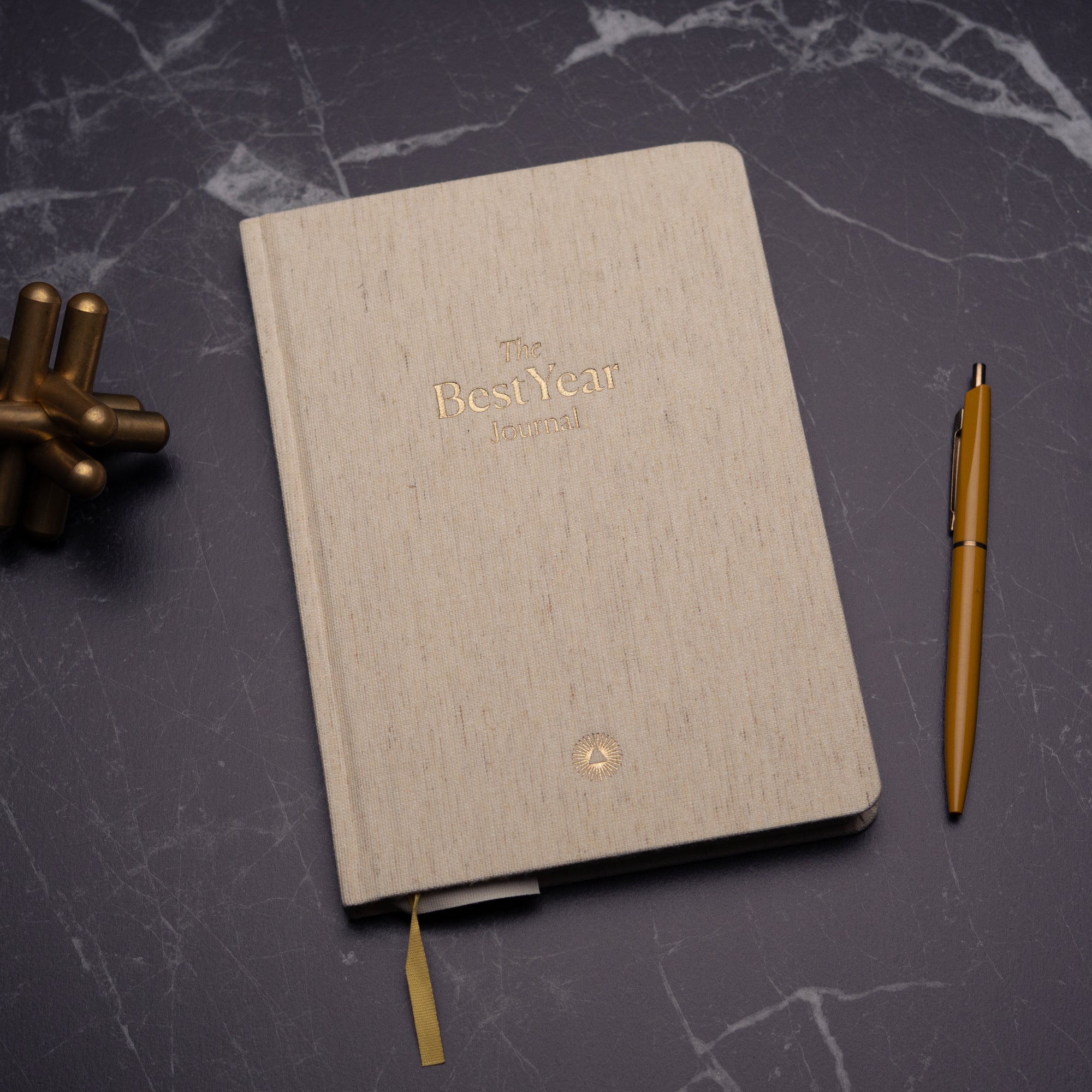 The Best Year Journal by Intelligent Change, a beige fabric-covered planner, rests closed on dark marble alongside a gold pen and metallic décor—ideal for mindfulness and intentional living.