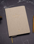 The Best Year Journal by Intelligent Change, a beige fabric-covered planner, rests closed on dark marble alongside a gold pen and metallic décor—ideal for mindfulness and intentional living.