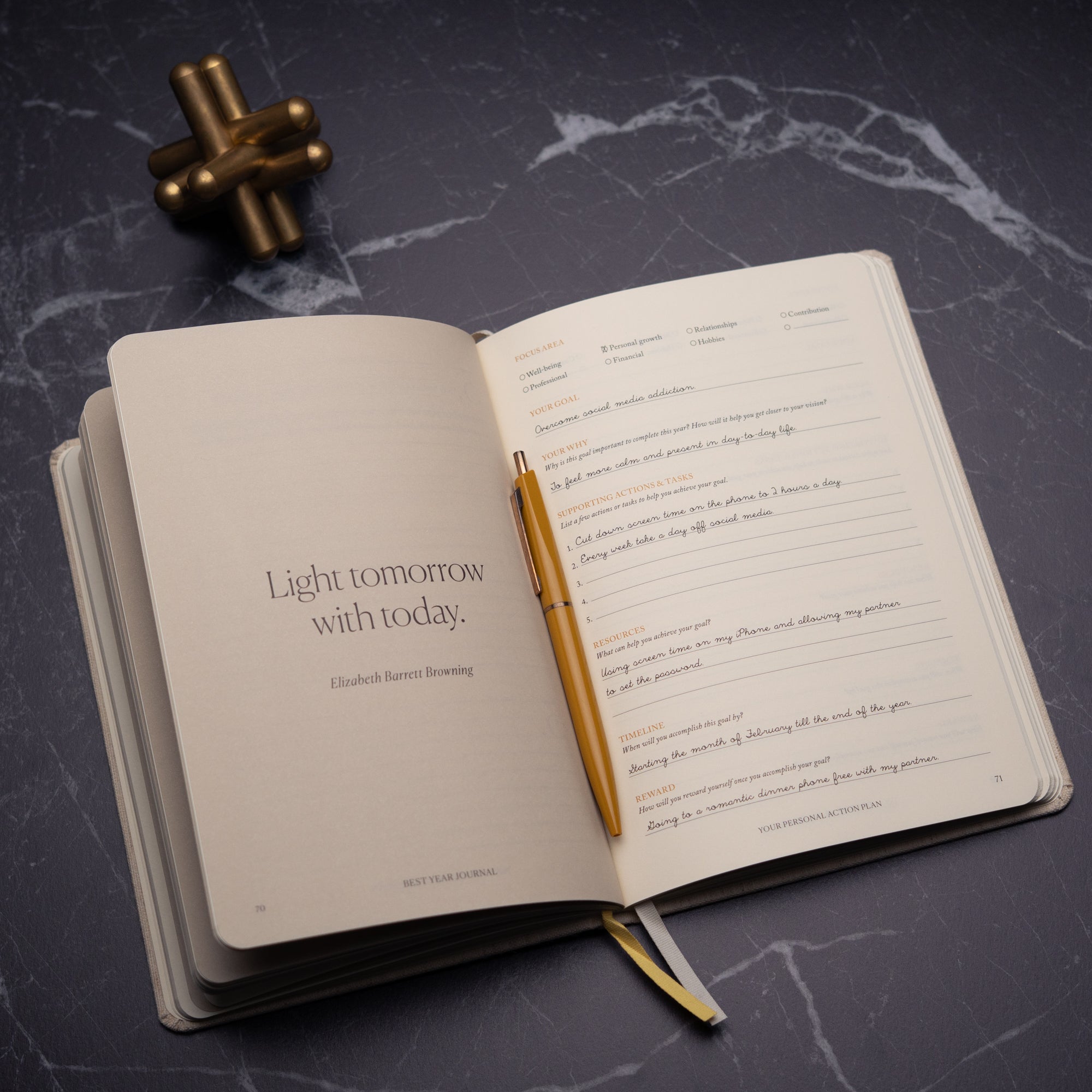 The Best Year Journal by Intelligent Change lies open on a dark marble surface with a gold pen, displaying the quote “Light tomorrow with today” by Elizabeth Barrett Browning. A gold desk ornament sits nearby, inspiring mindfulness and intention.