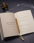The Best Year Journal by Intelligent Change lies open on a dark marble surface with a gold pen, displaying the quote “Light tomorrow with today” by Elizabeth Barrett Browning. A gold desk ornament sits nearby, inspiring mindfulness and intention.