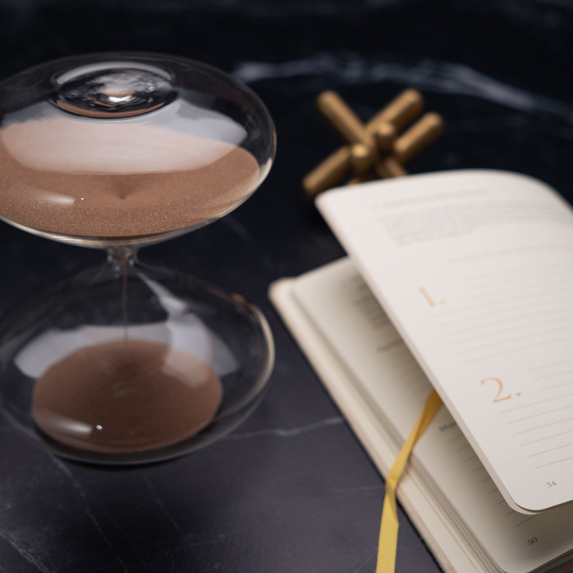 The Mindful Focus 30 Minute Hourglass by Intelligent Change, featuring brown sand, rests beside an open numbered journal with a yellow ribbon bookmark on a dark marble surface. A metallic object is blurred in the background.