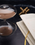 The Mindful Focus 30 Minute Hourglass by Intelligent Change, featuring brown sand, rests beside an open numbered journal with a yellow ribbon bookmark on a dark marble surface. A metallic object is blurred in the background.