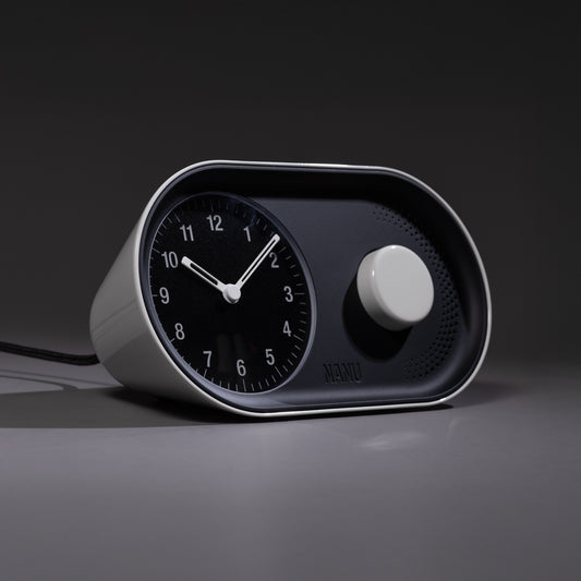 The Nanu Arc Alarm Clock features a modern black and white analog design with a circular dial and large knob, sitting on a smooth surface and casting a shadow in minimal lighting. Battery backup ensures uninterrupted timekeeping.