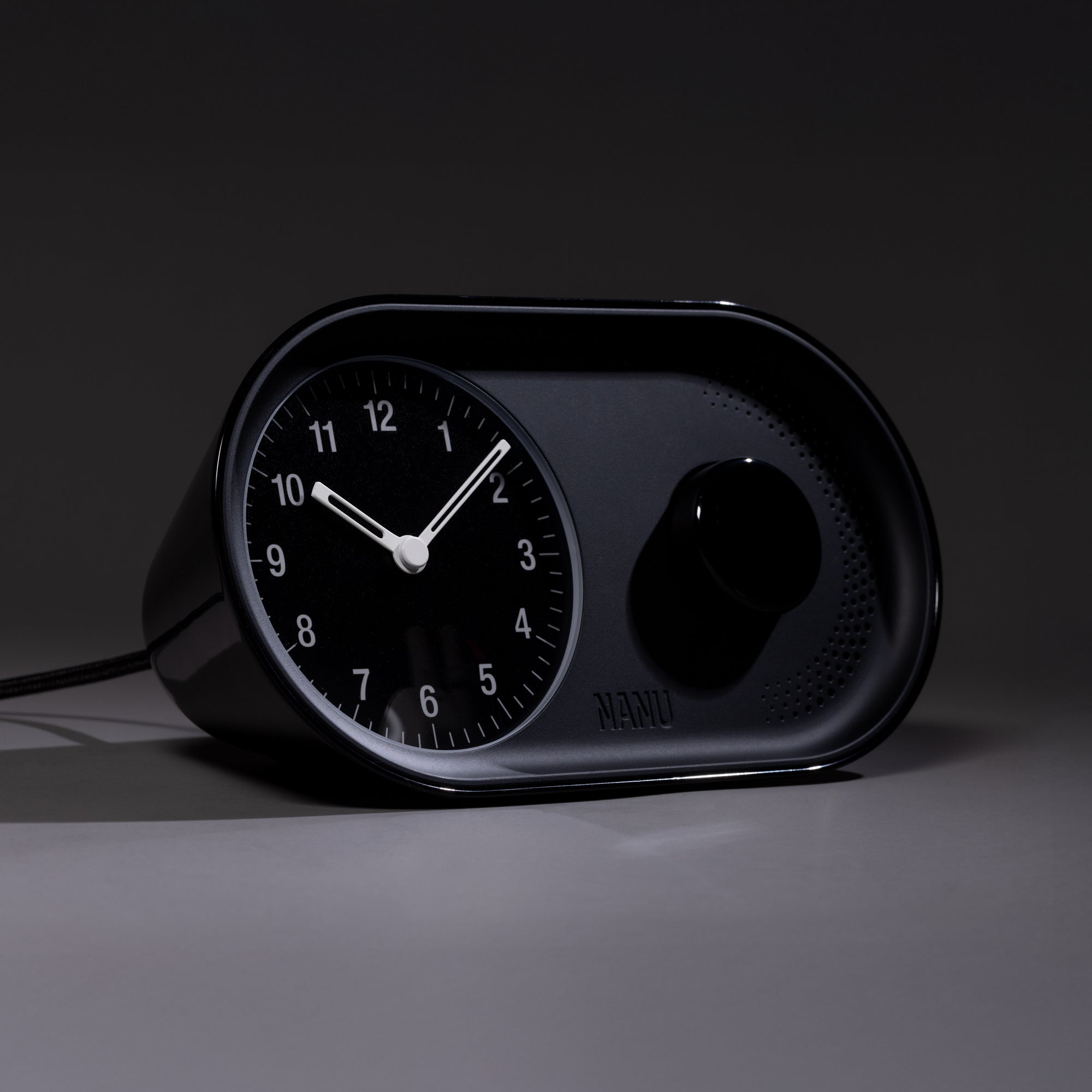 The Nanu Arc Alarm Clock, a modern black analog design with white hands and numbers, rests on a gray surface. It features minimalist style, battery backup, and a large circular knob beside its face.