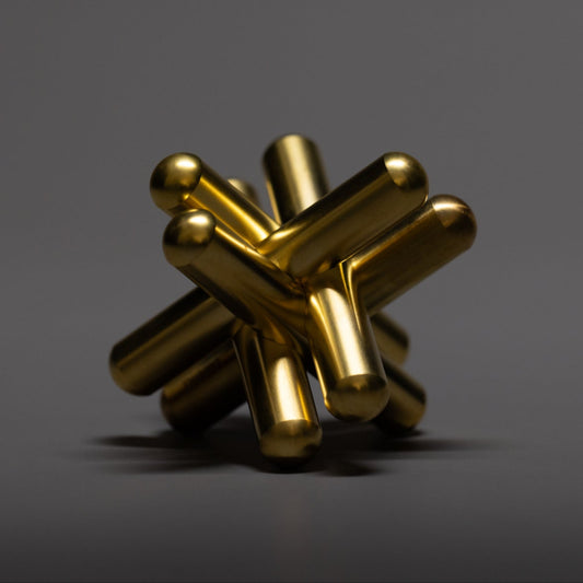 A metallic, gold-colored abstract sculpture with cylindrical rods intersecting at various angles, featuring a sleek modern design, set against a plain dark gray background.