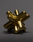 A metallic, gold-colored abstract sculpture with cylindrical rods intersecting at various angles, featuring a sleek modern design, set against a plain dark gray background.