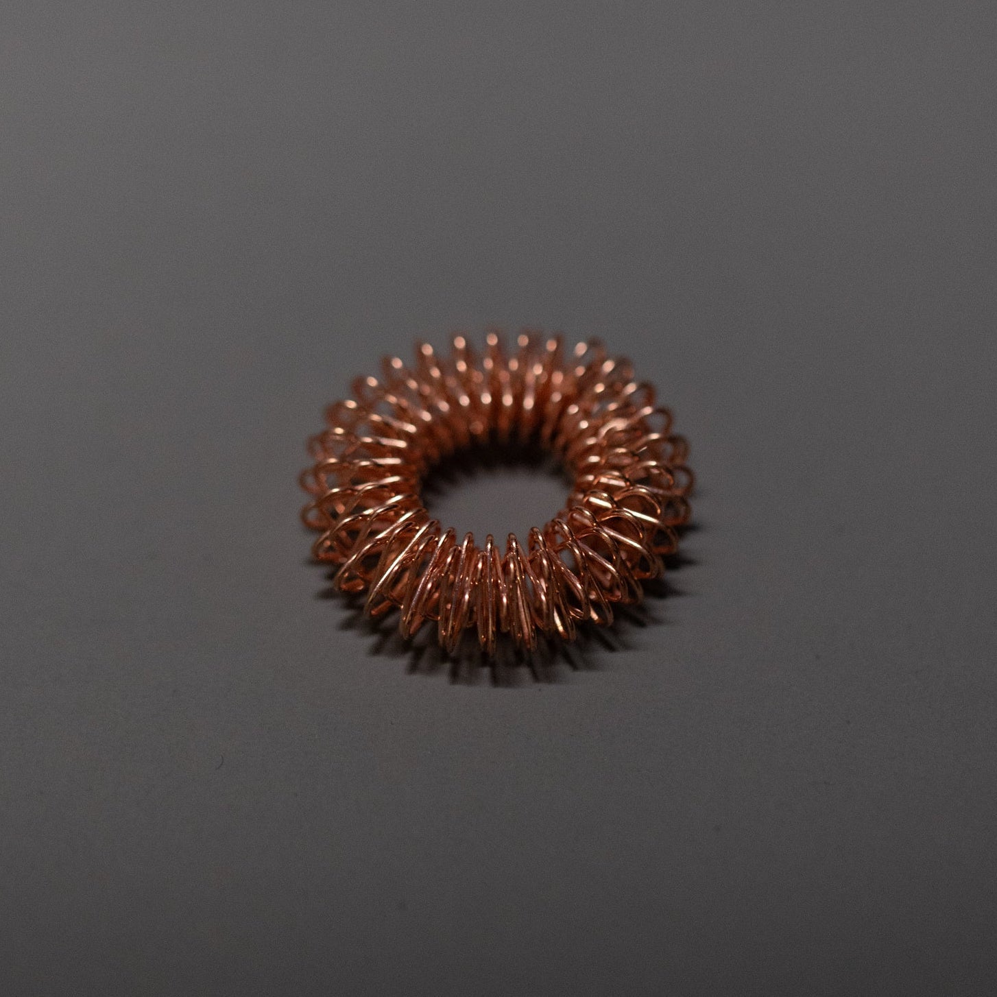 A small, circular copper wire coil is placed on a plain, dark gray surface. The tightly wound coil forms a donut-like shape, with evenly spaced loops creating an industrial, textured ring.