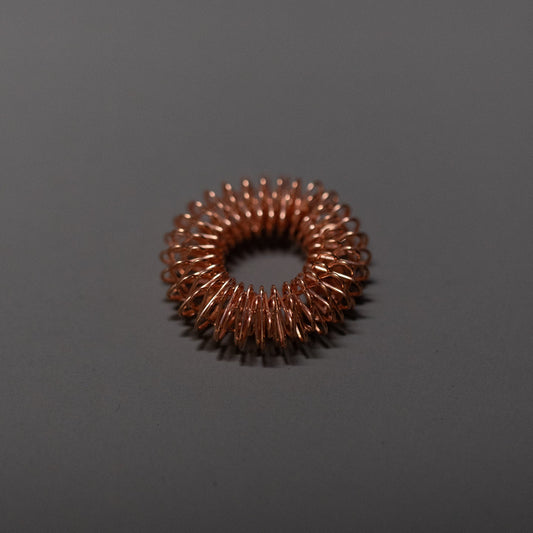 A small, circular copper wire coil is placed on a plain, dark gray surface. The tightly wound coil forms a donut-like shape, with evenly spaced loops creating an industrial, textured ring.