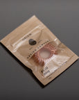 A small brown paper package labeled DESIGN COMPANY holds a pink, coiled ring-shaped accessory, possibly a hair tie, set against a plain gray background.