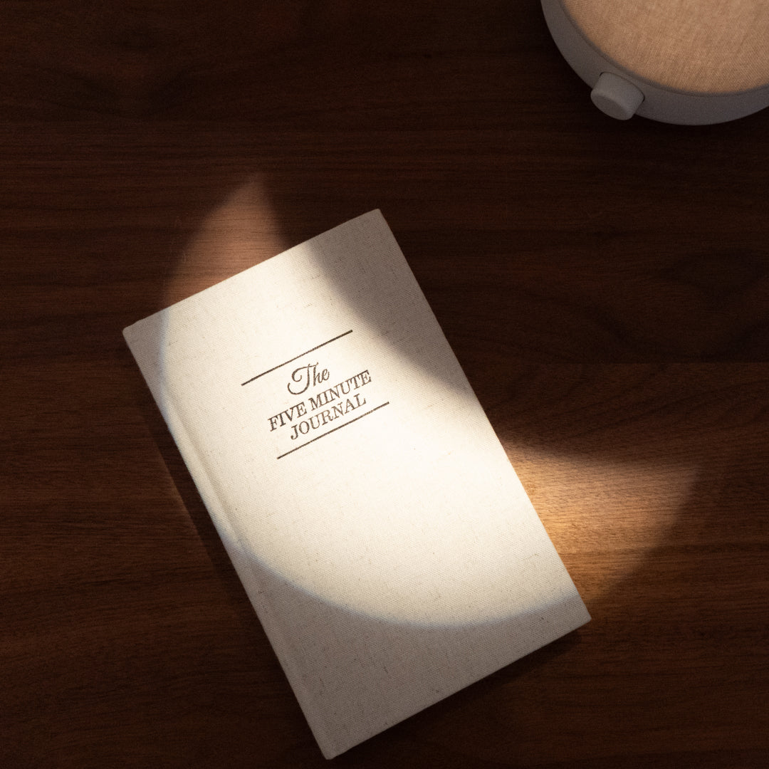 An undated Five Minute Journal by Intelligent Change rests on a wooden surface, partially lit by a circular light. It has a plain, light-colored cover with the title in bold black letters and includes 264 pages for six months of mindful reflection.