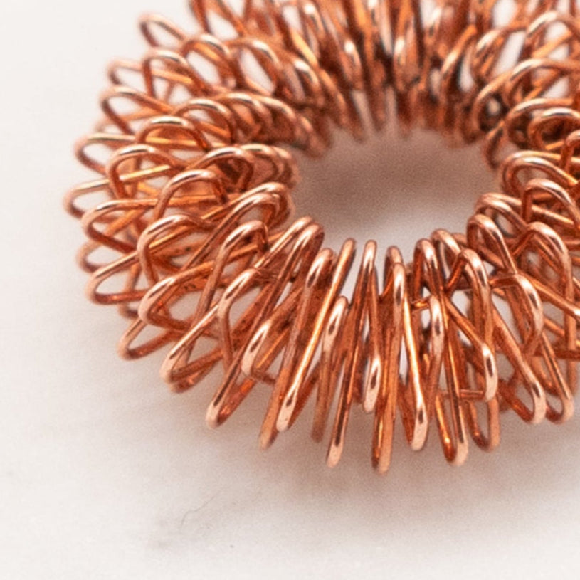The copper-colored FL·O Ring Finger Massager by Orijin, a coiled metal ring, elegantly rests on a white marble surface, showcasing its tightly wound precision and refined form.