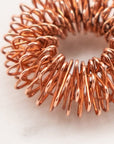 The copper-colored FL·O Ring Finger Massager by Orijin, a coiled metal ring, elegantly rests on a white marble surface, showcasing its tightly wound precision and refined form.