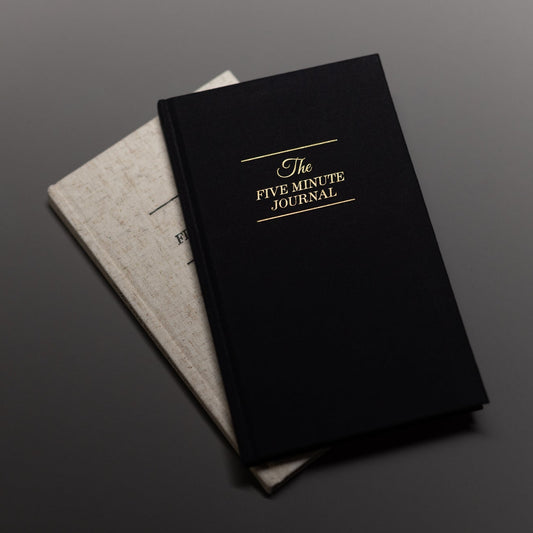 Two journals are stacked on a dark surface. The top journal, The Five Minute Journal, features elegant gold text on a black cover. A light beige notebook rests partially visible beneath it, creating a minimalist scene.