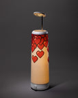A cylindrical lamp with a white base and top, decorated with a pattern of red pixelated hearts. The lamp emits a warm light and has a modern, minimalist design against a dark gray background.