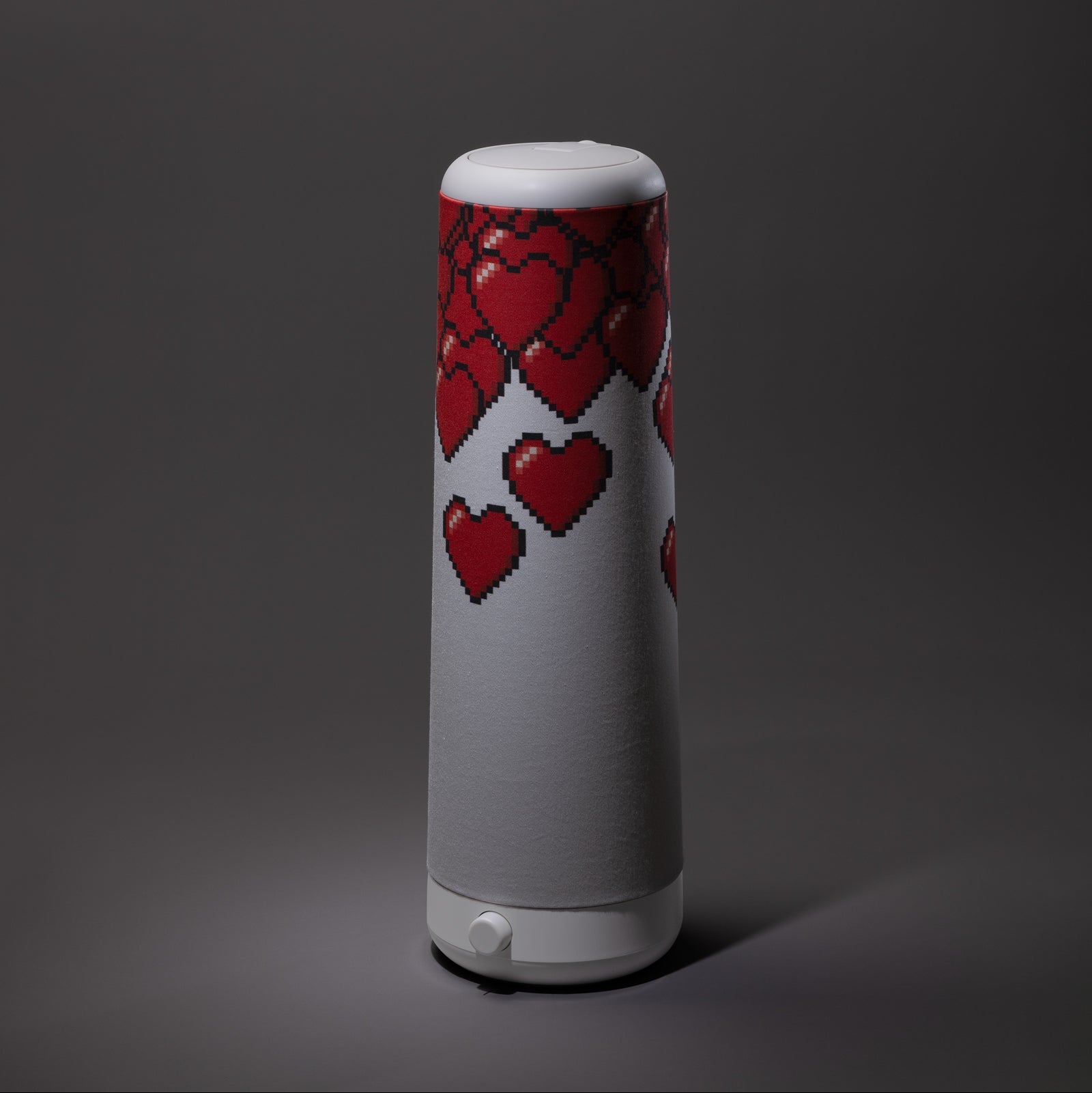 A tall, cylindrical white table lamp decorated with pixelated red hearts on its upper half, set against a dark gray background.