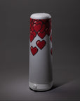 A tall, cylindrical white table lamp decorated with pixelated red hearts on its upper half, set against a dark gray background.