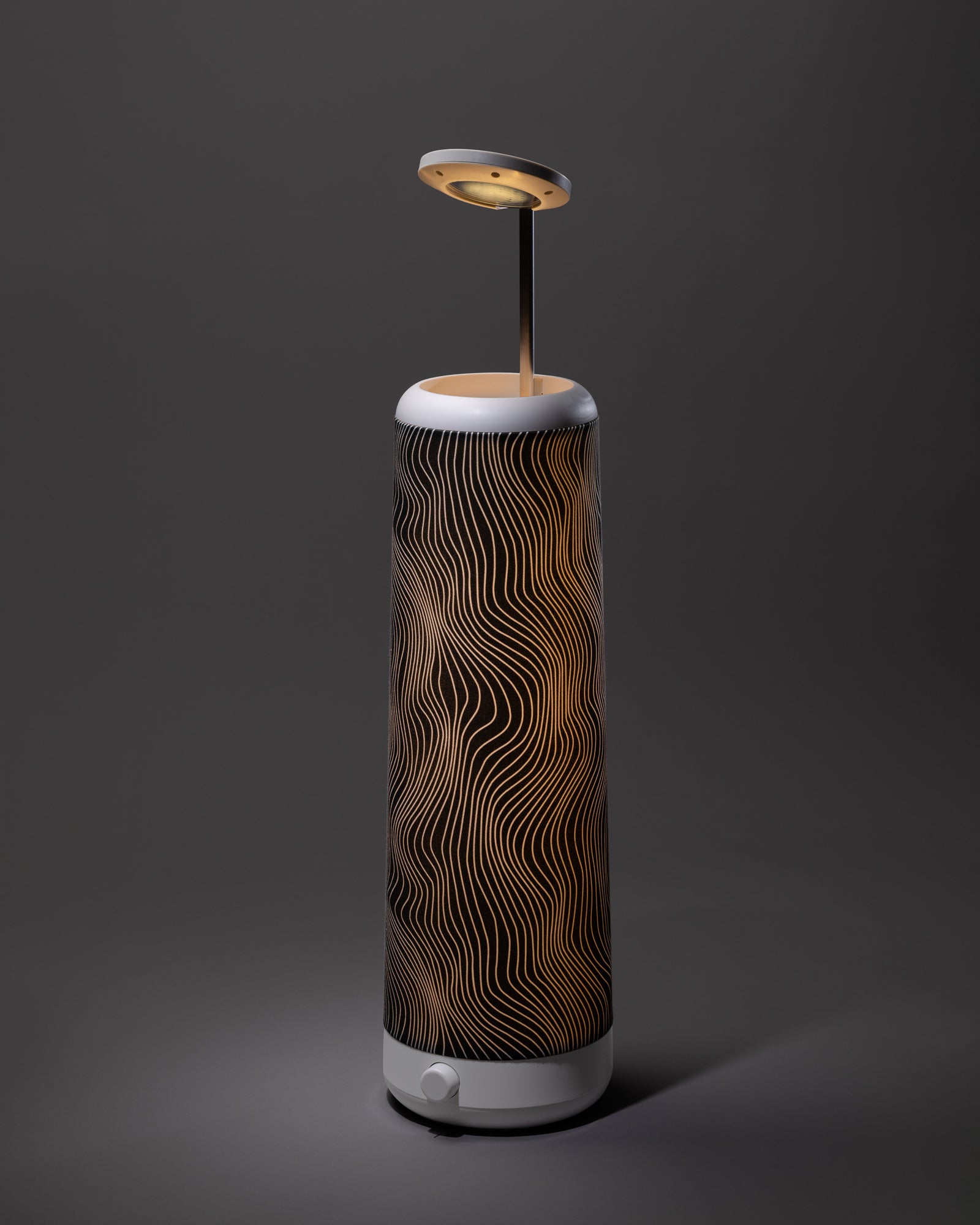 A modern table lamp with a cylindrical base featuring wavy black and gold lines, a round white knob at the bottom, and a flat circular light source suspended above the base on a thin metal rod.