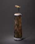 A modern table lamp with a cylindrical base featuring wavy black and gold lines, a round white knob at the bottom, and a flat circular light source suspended above the base on a thin metal rod.