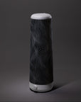A tall, cylindrical table lamp with a white base and top, featuring a black body covered in wavy, white line patterns, set against a plain gray background.
