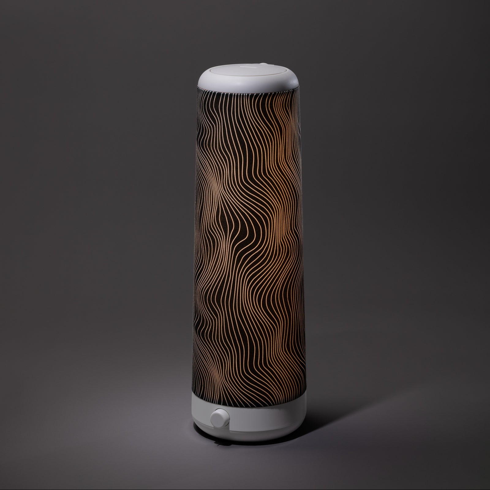 A tall, cylindrical lamp with a white base and top, featuring a black and gold wavy line pattern on its shade, is illuminated against a plain grey background.