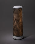 A tall, cylindrical lamp with a white base and top, featuring a black and gold wavy line pattern on its shade, is illuminated against a plain grey background.