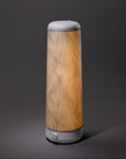 A modern, cylindrical lamp with a wavy line pattern on its shade emits a warm yellow light, standing on a plain gray surface against a dark gray background.