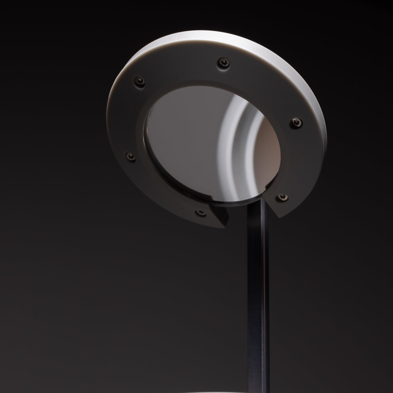 A close-up of the NS01 Personal Task Lamp by Nightside, featuring a round gray shade and a circular metallic accent, hand assembled and shown against a dark background.