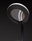 A close-up of the NS01 Personal Task Lamp by Nightside, featuring a round gray shade and a circular metallic accent, hand assembled and shown against a dark background.