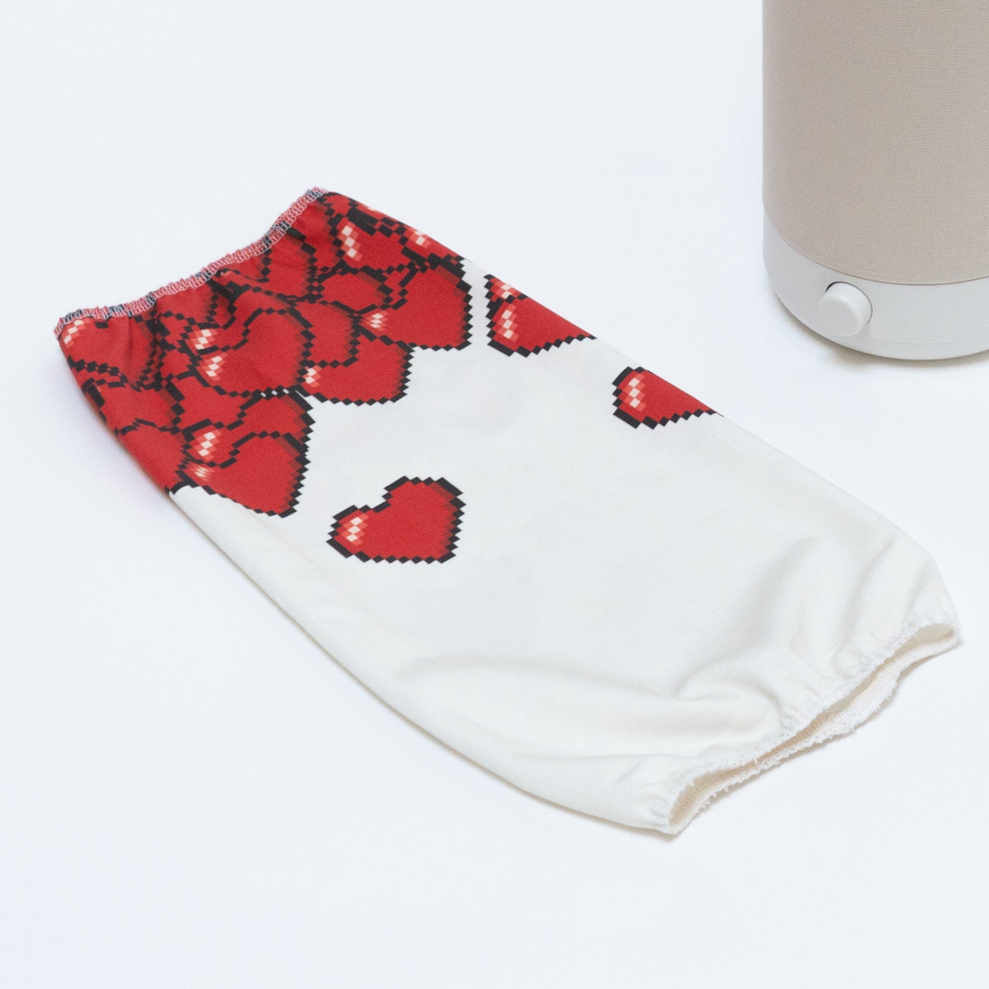 The 8-bit Hearts Cotton Shade, featuring red pixelated heart designs on white fabric, is displayed on a white surface next to the light beige base of a Nightside NS01 task lamp, awaiting its interchangeable shade.