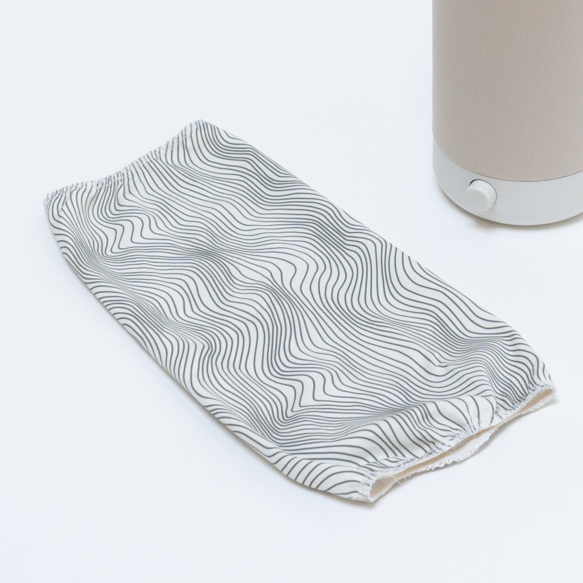 A white, elongated fabric handle cover with black wavy lines lies on a light surface. To its right, an object resembling the Electric Flow Field White Cotton Shade from Nightside is partially visible, with hints of interchangeable shades for versatile decor thanks to its beige base and white knob.