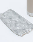 A white, elongated fabric handle cover with black wavy lines lies on a light surface. To its right, an object resembling the Electric Flow Field White Cotton Shade from Nightside is partially visible, with hints of interchangeable shades for versatile decor thanks to its beige base and white knob.