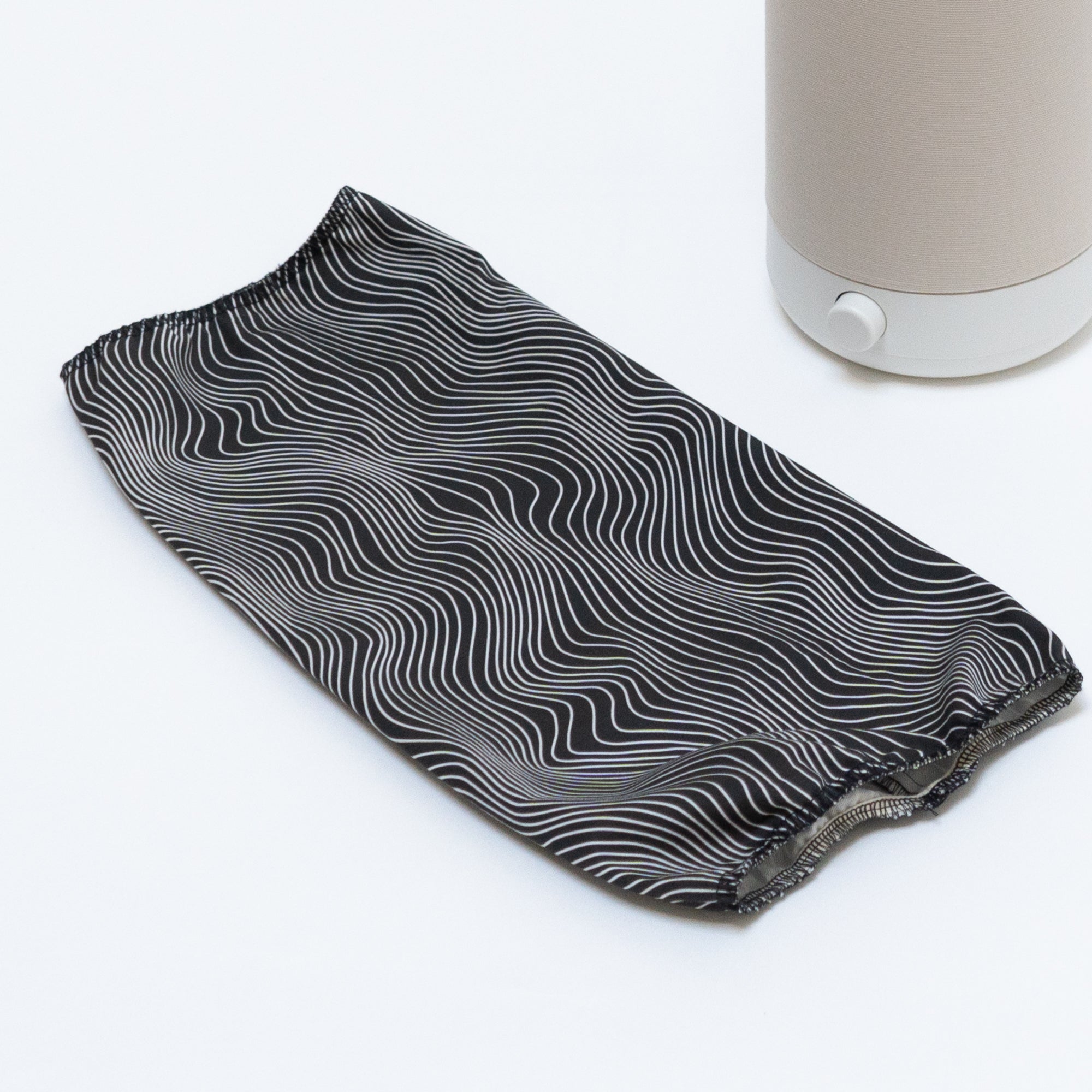 On a white surface rests a Nightside Electric Flow Field shade, a black fabric sleeve adorned with wavy white lines. Nearby, the cylindrical beige Nightside NS01 task lamp with its white base hints at its interchangeable cotton shades.