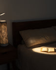 A dimly lit bedroom features a closed book on the bed, softly illuminated by a Nightside Electric Flow Field lamp with its wavy pattern. The cozy atmosphere is enhanced by the wooden headboard, nightstand, and blackout shades.