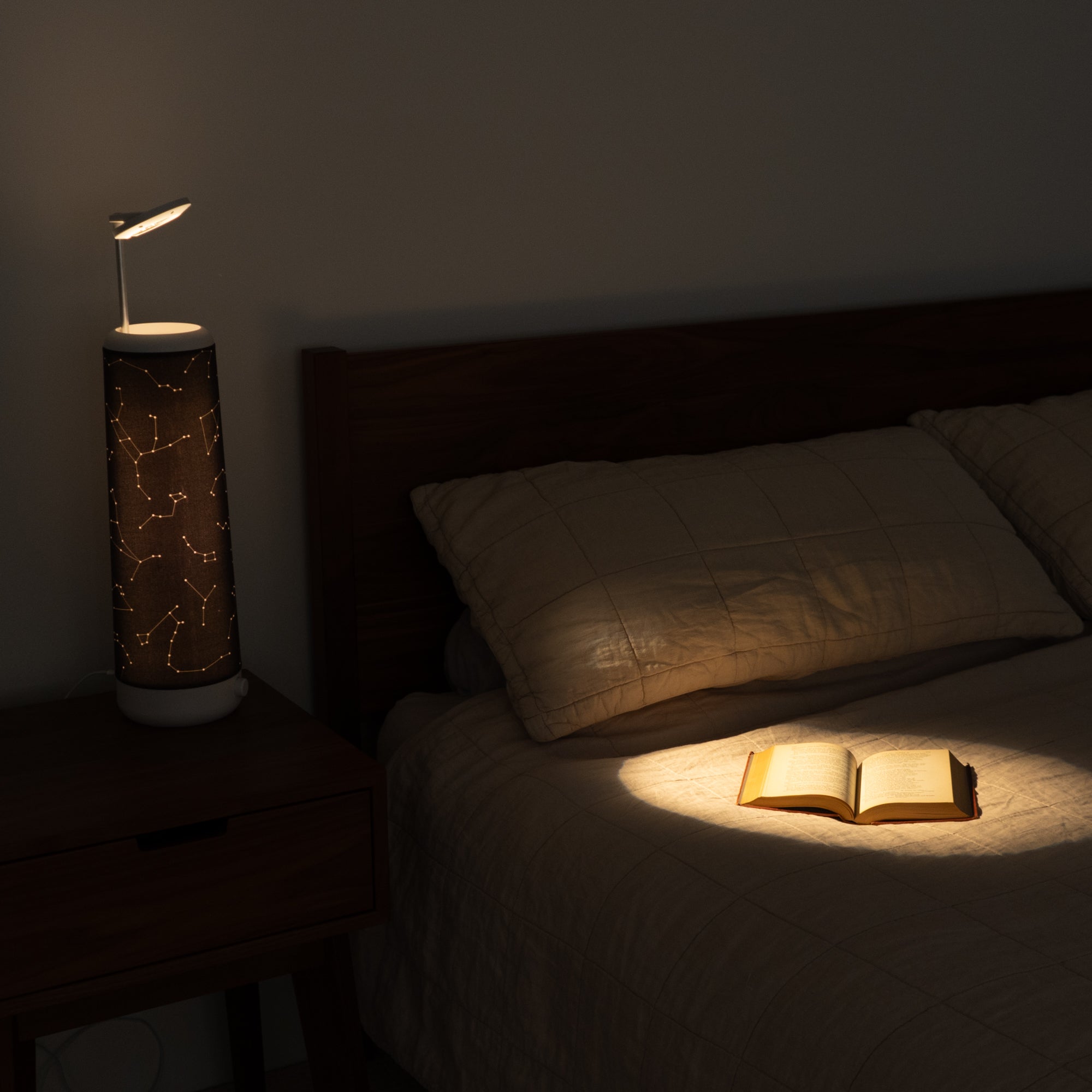 A cozy bedroom features a bed with beige plaid bedding and an open book under the constellation-themed lamp by Nightside on a wooden nightstand. Dim lighting, coupled with interchangeable fabric shades from the Starlight Cotton Blackout Shade collection, creates a warm ambiance.