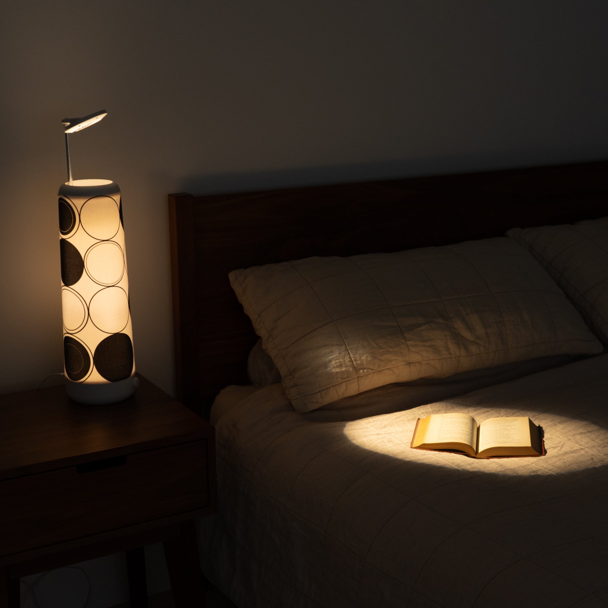 The bedroom features a bed with a beige quilt, softly lit by the Nightside lamps Concentric Circles Cotton Shade. Warm light highlights an open book on the bed, while blackout shades keep the room dim and cozy.