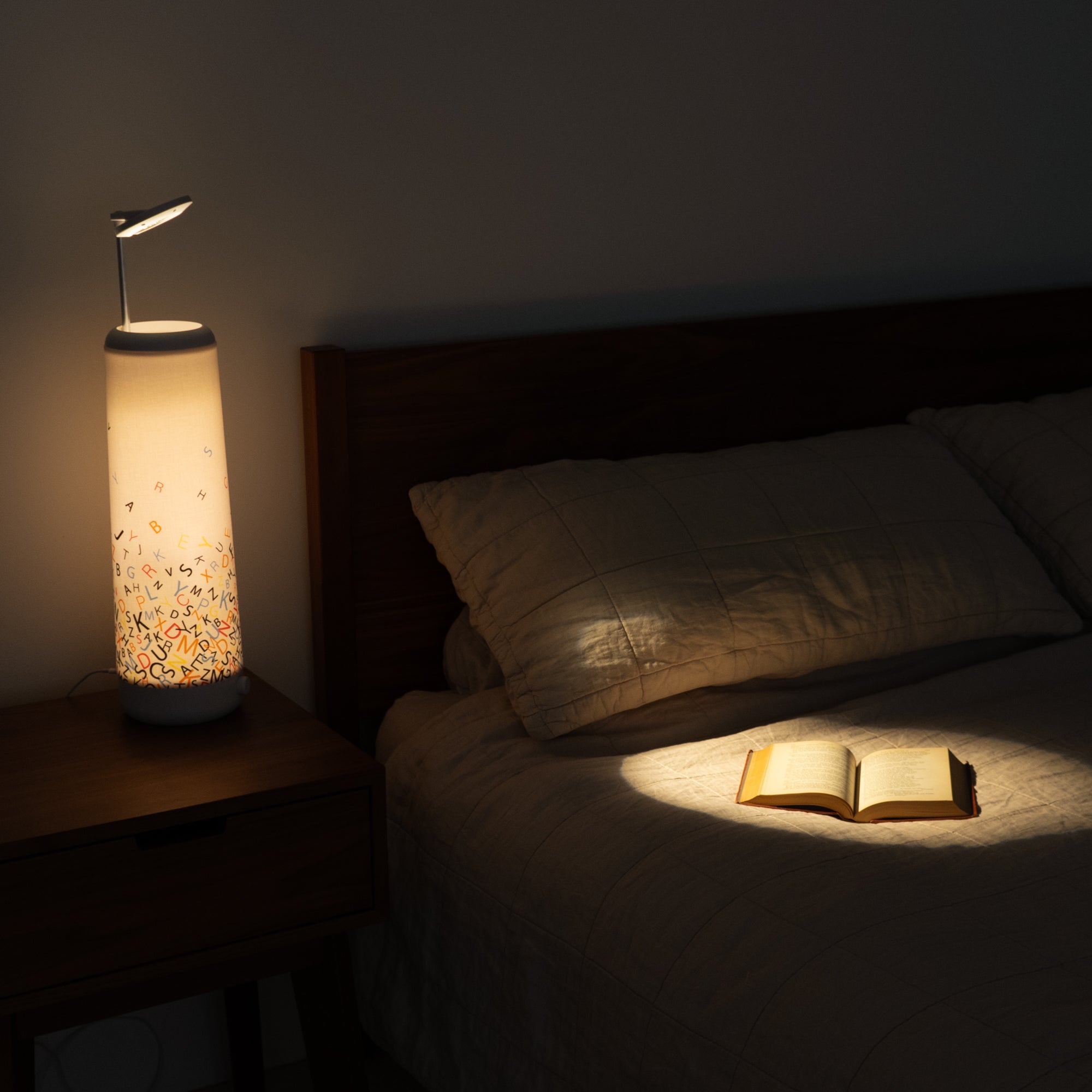 A cozy bedroom scene showcases a Nightside lamp with the Alphabet Cascade Cotton Shade, illuminating an open book on a neatly made bed. The warm glow highlights the pillow and partially lights up the wooden headboard and nightstand, with fabric shades offering versatile style.