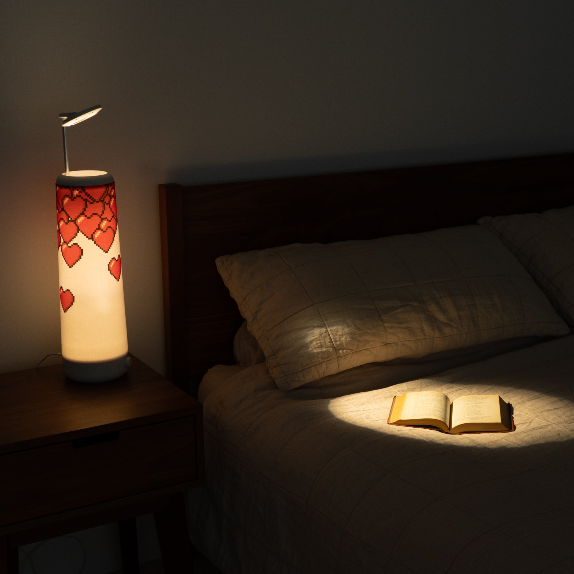 A cozy bedroom with a dimly lit Nightside lamp featuring an 8-bit Hearts Cotton Shade on a bedside table. An open book on the bed is illuminated by a focused spotlight against a quilted cover, and the lamps interchangeable fabric shades add personal flair to the tranquil setting.