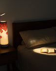 A cozy bedroom with a dimly lit Nightside lamp featuring an 8-bit Hearts Cotton Shade on a bedside table. An open book on the bed is illuminated by a focused spotlight against a quilted cover, and the lamps interchangeable fabric shades add personal flair to the tranquil setting.
