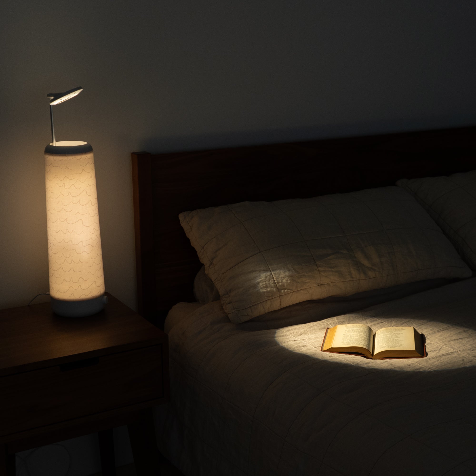 A cozy bedroom features a dimly lit Nightside lamp with a Cursive Lines Cotton Shade, illuminating an open book on the neatly made bed. The interchangeable fabric shades cast a soft glow, creating a warm and inviting atmosphere against the dark background.