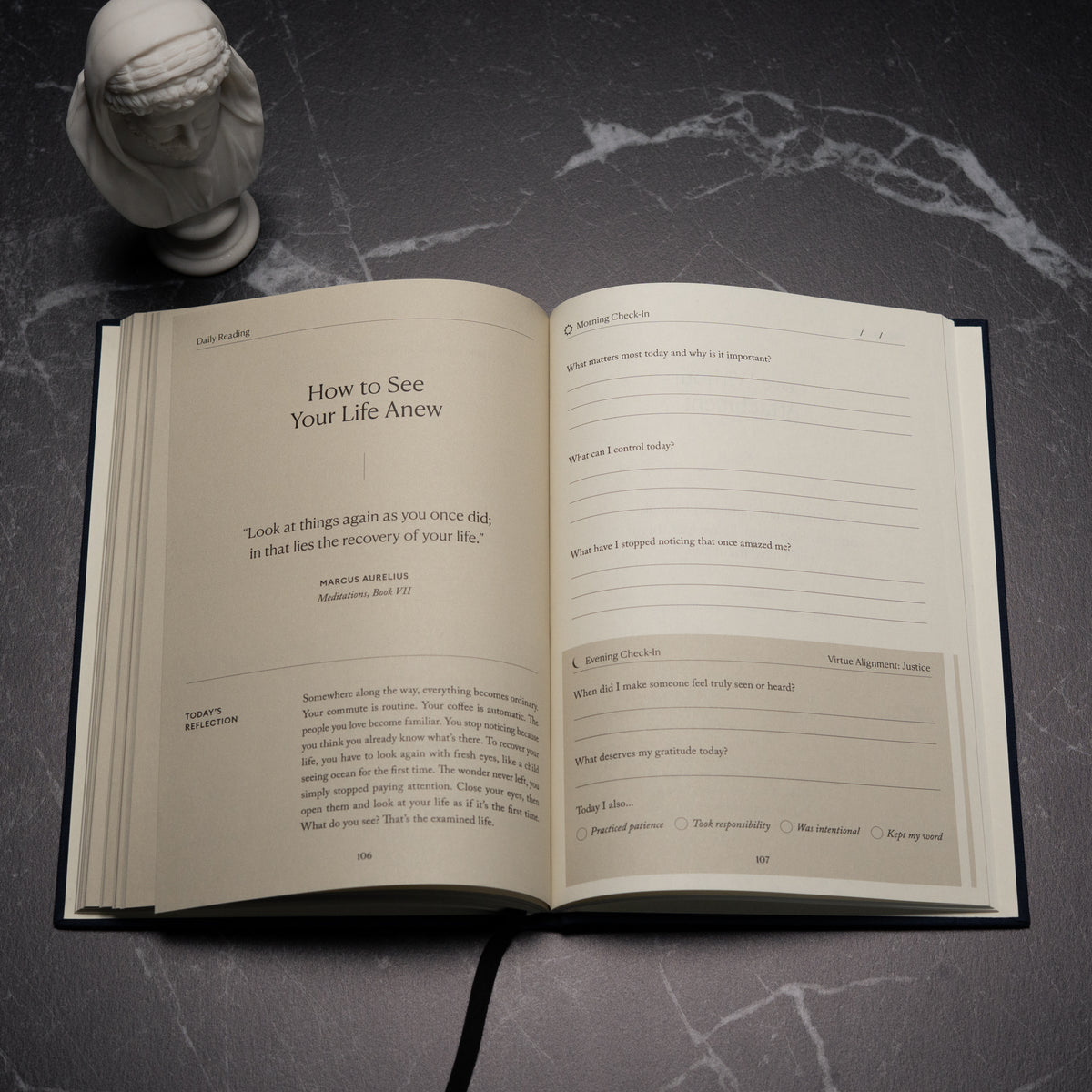 An open copy of The Stoic Journal by Intelligent Change rests on a dark surface beside a small white bust. The visible pages present daily reflection prompts and a Marcus Aurelius quote about seeing life anew, encouraging you to embrace Stoic philosophy in your everyday practice.
