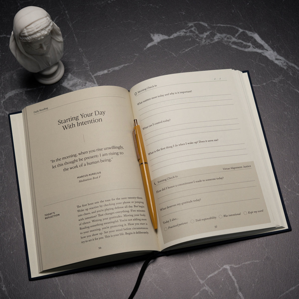 An open copy of The Stoic Journal by Intelligent Change, accompanied by a gold pen, rests on a dark surface beside a small white bust statue. The pages of the journal offer daily prompts and reflections inspired by Stoic philosophy, encouraging mindfulness and intentional living.
