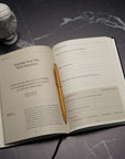An open copy of The Stoic Journal by Intelligent Change, accompanied by a gold pen, rests on a dark surface beside a small white bust statue. The pages of the journal offer daily prompts and reflections inspired by Stoic philosophy, encouraging mindfulness and intentional living.