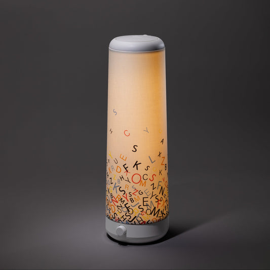 The Nightside Alphabet Cascade Cotton Shade is a modern cylindrical lamp with interchangeable fabric shades and a soft warm glow, featuring colorful scattered alphabet letters near its base against a dark grey background.