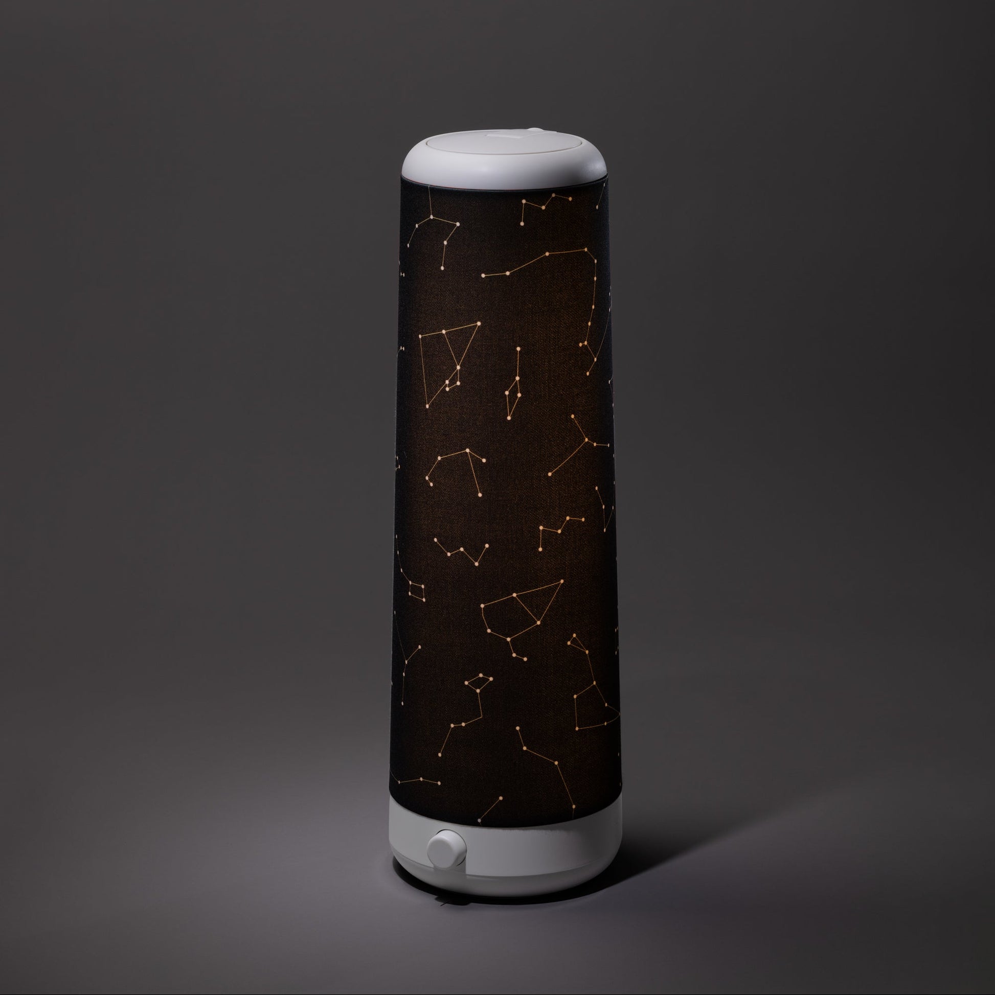 A cylindrical Nightside lamp with a Starlight Cotton Blackout Shade, featuring golden constellation patterns, stands on a plain gray surface against a gray background.
